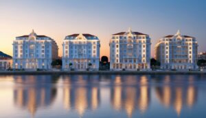 Four ornate buildings with red roofs reflected in calm water at sunset